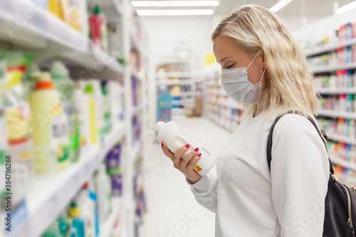 Young woman wearing medical mask chooses home cleaning product in supermarket. Coronavirus pandemic.