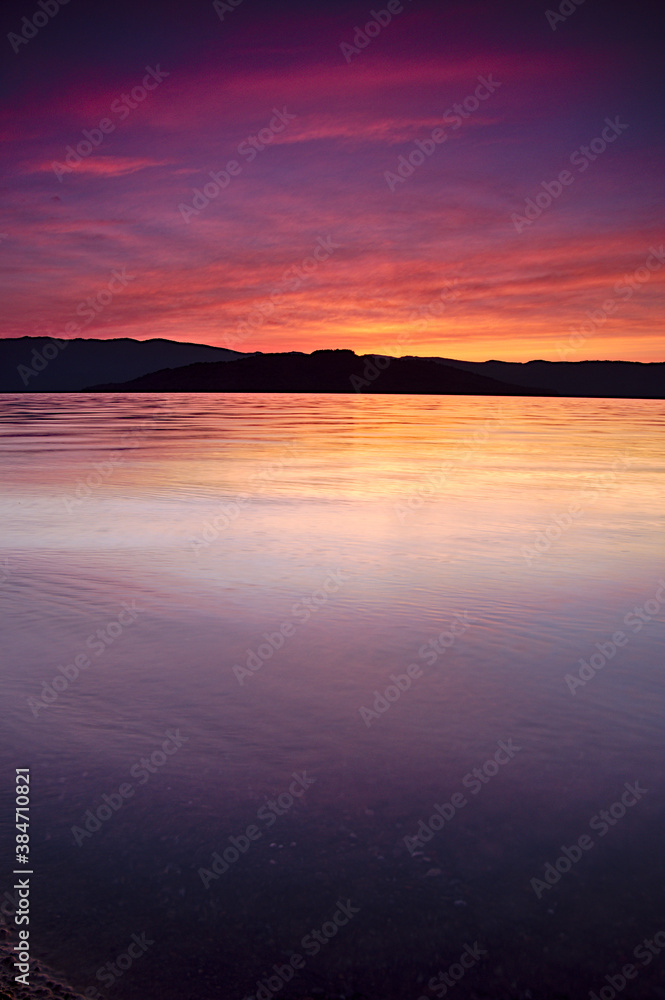 夕暮れの湖。穏やかな波の水面に映る空の色。遠くの山のシルエット。