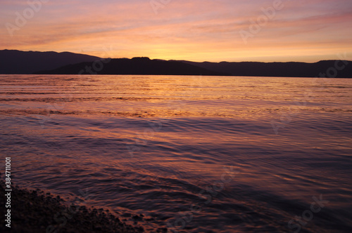 夕暮れの湖。穏やかな波の水面に映る空の色。遠くの山のシルエット。