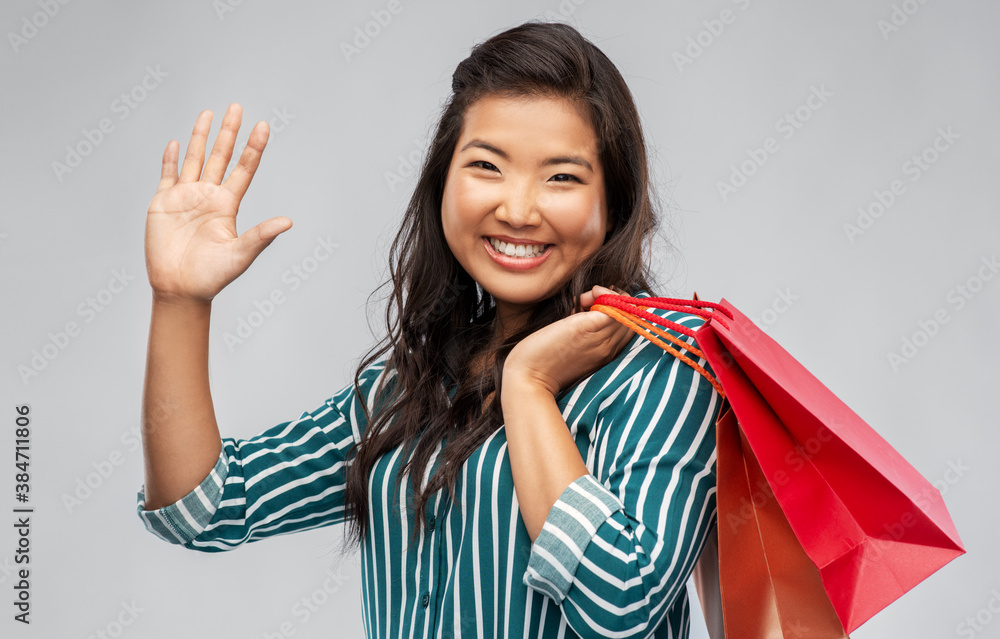 sale, outlet and consumerism concept - happy asian young woman with shopping bags waving hand over grey background sale, outlet and consumerism concept - happy asian young woman with shopping bags waving hand over grey background