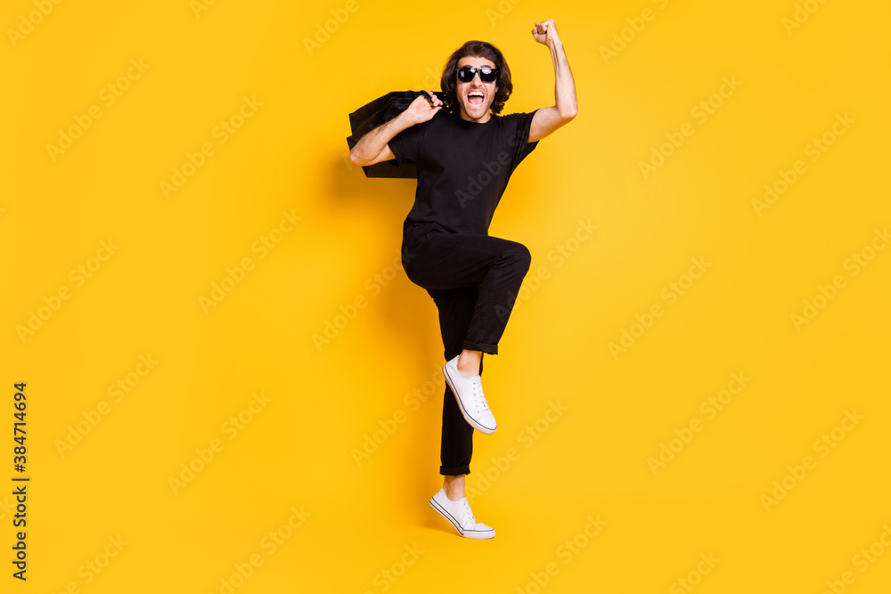 Full length photo of young man hold shopping packs open mouth raise hand wear black t-shirt pants white sneakers specs isolated yellow color background