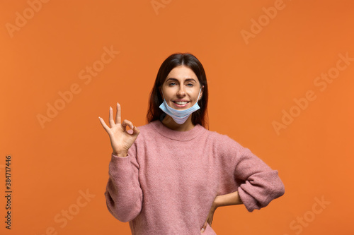 Attractive woman put medical mask on the chin saying okay showing ok or perfect gesture. Pleased give positive reply like excellent job. wears in pink sweater isolated. Tired of coronavirus.