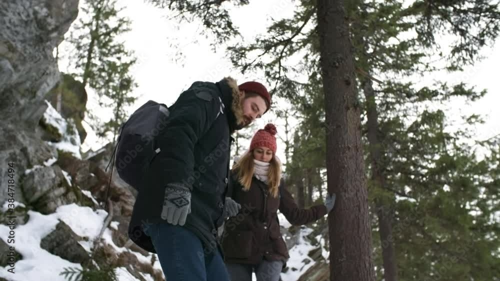 Tracking shot of happy young couple with backpacks holding hands and descending mountain while hiking in winter Tracking shot of happy young couple with backpacks holding hands and descending mountain while hiking in winter