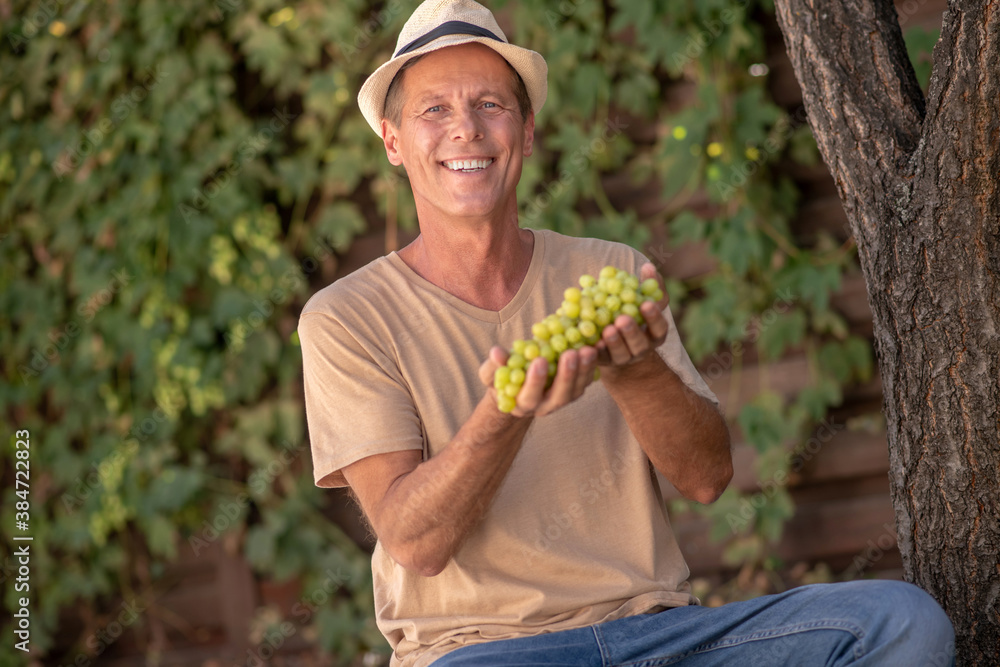 Smiling male in straw hat holding cluster of green grapes