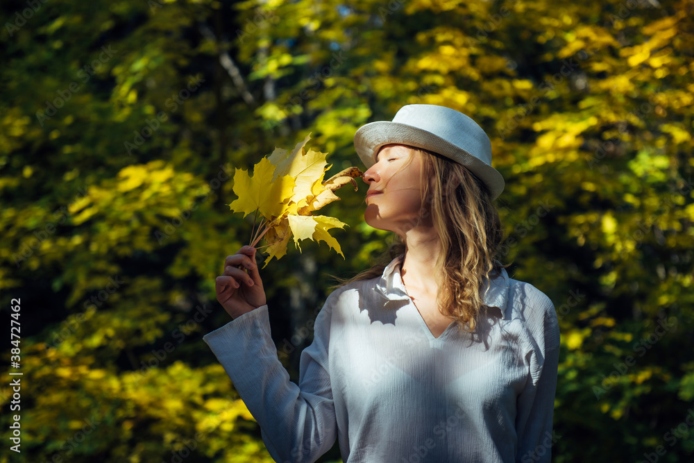 Elegant young blonde in white shirt and hat in autumn park on sunny day. Beautiful happy smiling woman held a bouquet of yellow maple leaves to her face. People in outdoor leisure activity.