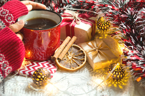 A woman's hand in a warm sweater holds a red mug with a hot drink on a table with Christmas decorations. New year's atmosphere, cinnamon sticks and a slice of dried orange, gifts, garland and tinsel