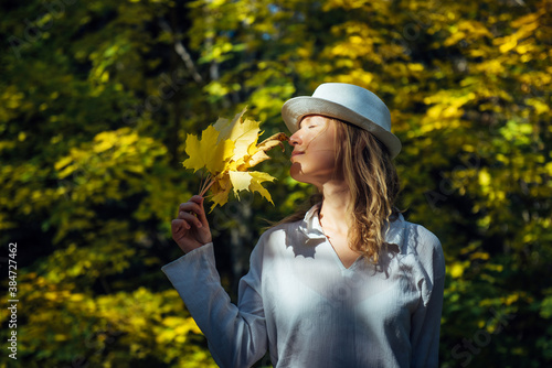 Elegant young blonde in white shirt and hat in autumn park on sunny day. Beautiful happy smiling woman held a bouquet of yellow maple leaves to her face. People in outdoor leisure activity.