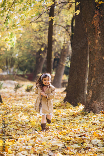 Happy and beautiful little girl in a beige coat runs holding a maple leaf in the autumn park