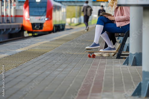 Legs of a girl in white socks and sneakers  leaning on a skateboard. A girl sits on a train station bench against the background of an arriving train on a sunny summer day. Travel light.