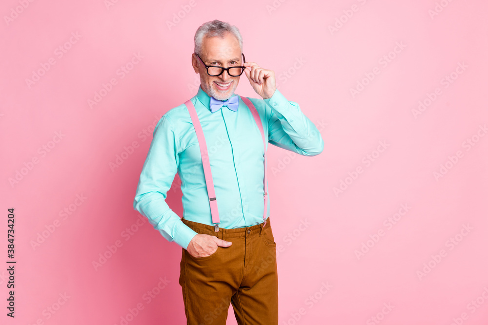 Photo portrait of happy smiling elder man touching eyeglasses wearing bright shirt bowtie suspenders brown trousers isolated on pink color background Photo portrait of happy smiling elder man touching eyeglasses wearing bright shirt bowtie suspenders brown trousers isolated on pink color background