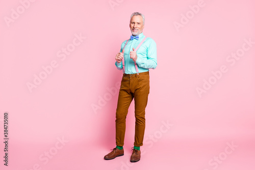 Full length body size portrait of smiling cheerful old gentleman wearing bowtie pants touching suspenders isolated on pink color background