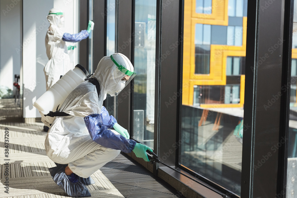 Side view full length portrait of two female workers wearing hazmat suits disinfecting windows in office building, copy space Side view full length portrait of two female workers wearing hazmat suits disinfecting windows in office building, copy space