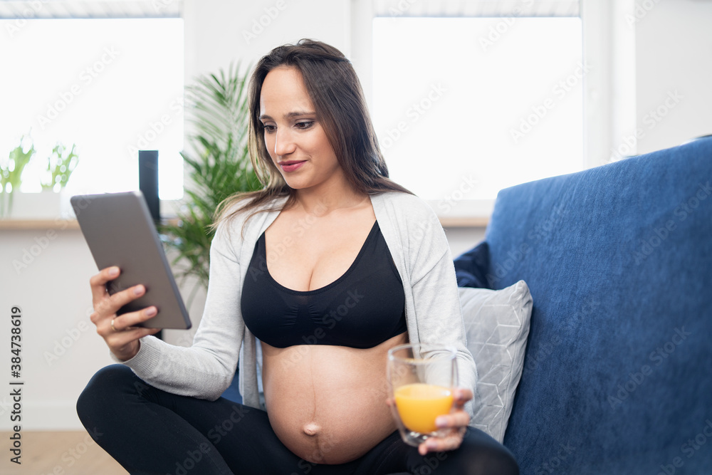 Smiling pregnant brunettre woman with a glass of orange juice is sitting on a sofa in the living room and reading articles on tablet. She is wearing sports clothes. Smiling pregnant brunettre woman with a glass of orange juice is sitting on a sofa in the living room and reading articles on tablet. She is wearing sports clothes.