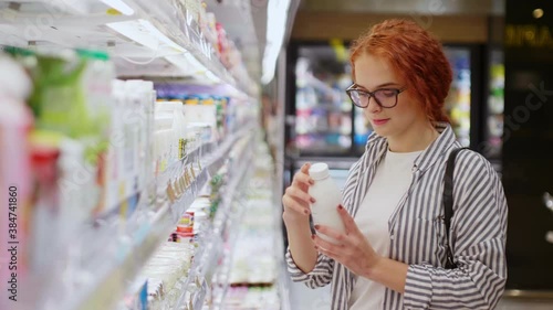 Attractive redhead girl in eyeglasses dressed in casual wear thoughtfully choosing yogurt in modern grocery store