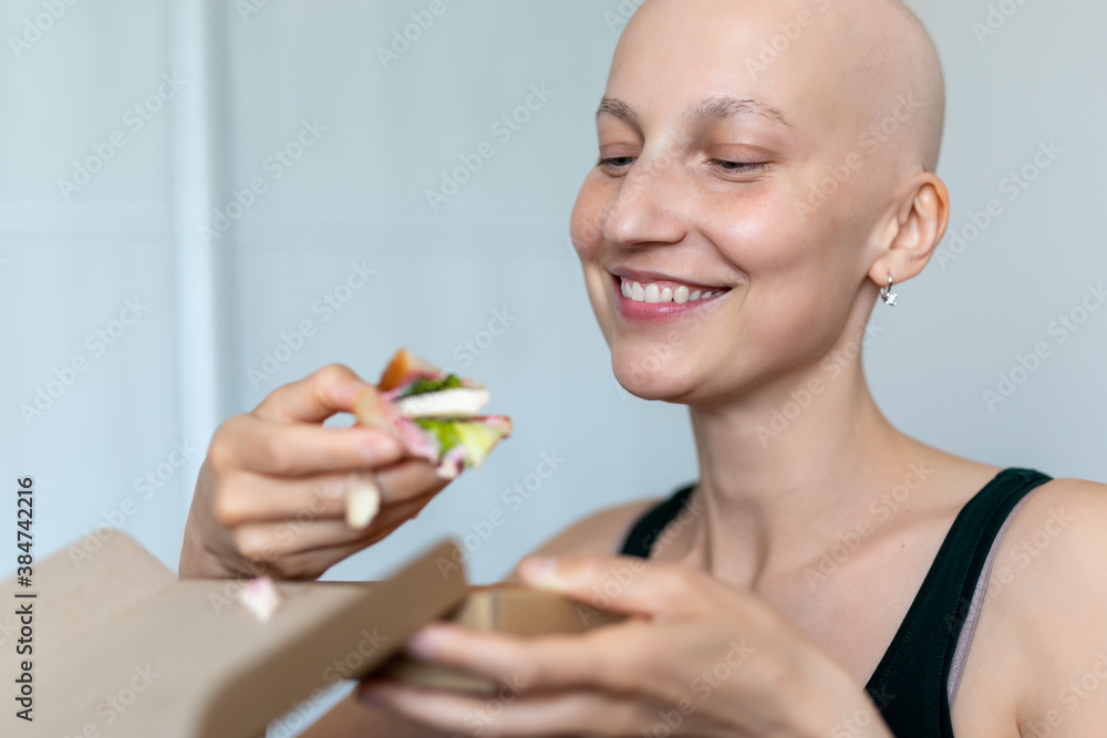 Young adult beautiful caucasian bald shaved hair woman portrait enjoy eating sandwich for lunch at home indoor. Breast cancer survivor person nutrition healthy diet awareness. Chemoterapy alopecia Young adult beautiful caucasian bald shaved hair woman portrait enjoy eating sandwich for lunch at home indoor. Breast cancer survivor person nutrition healthy diet awareness. Chemoterapy alopecia