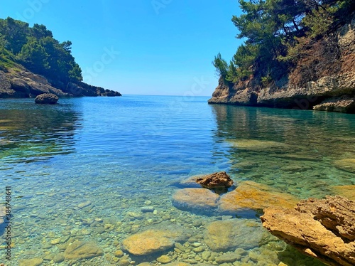 tropical island with pebble beach and crystal clear turquoise calm sea water surrounded pine tree coniferous forest over blue heaven, Mediterranean rocky coast Montenegro, summer vacation and holidays