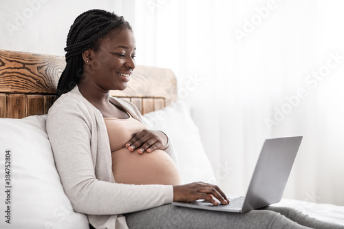 Charming pregnant woman relaxing with her laptop, browsing or chatting with friends online, sitting on bed, panorama with empty space