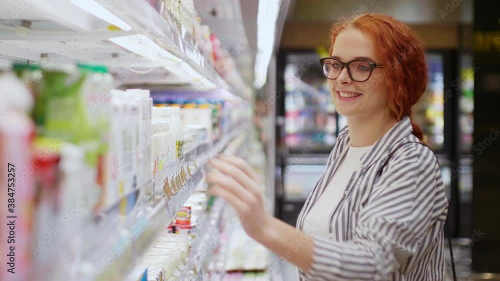 Pretty smiling redhead girl in eyeglasses dressed casual wear choosing yogurt in modern grocery store. Supermarket concept Pretty smiling redhead girl in eyeglasses dressed casual wear choosing yogurt in modern grocery store. Supermarket concept