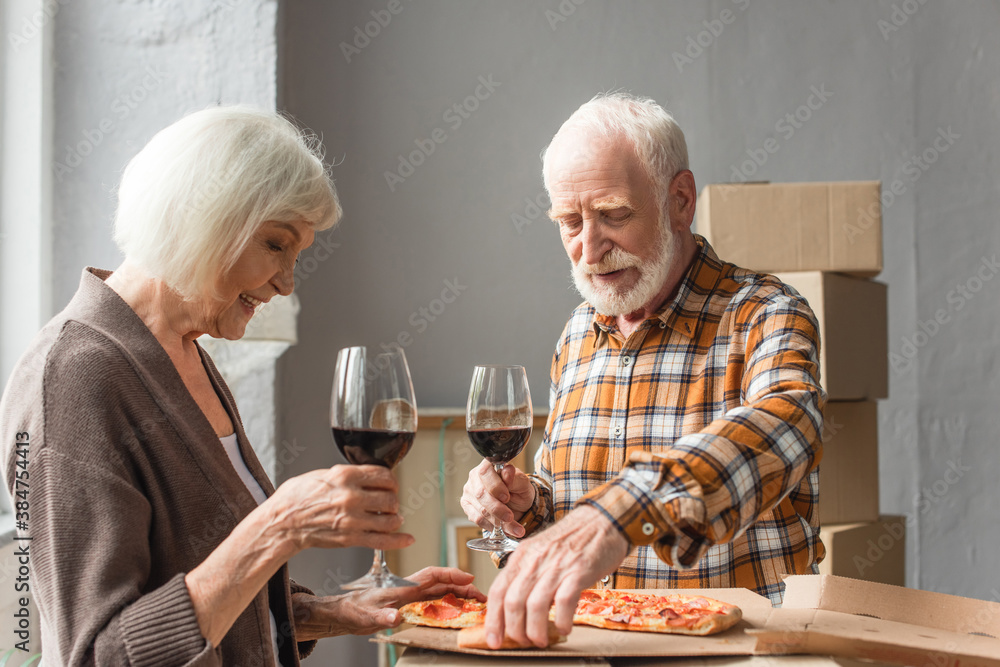 senior couple holding glasses of wine and man taking piece of pizza in new house