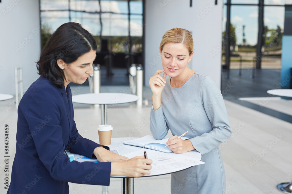 Fototapeta premium Waist up portrait of two successful businesswomen discussing deal while standing by cafe table in airport or office building