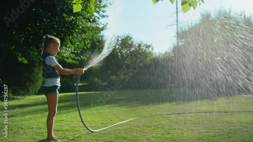 adorable little girl playing with a garden hose on hot and sunny summer evening, child watering green grass