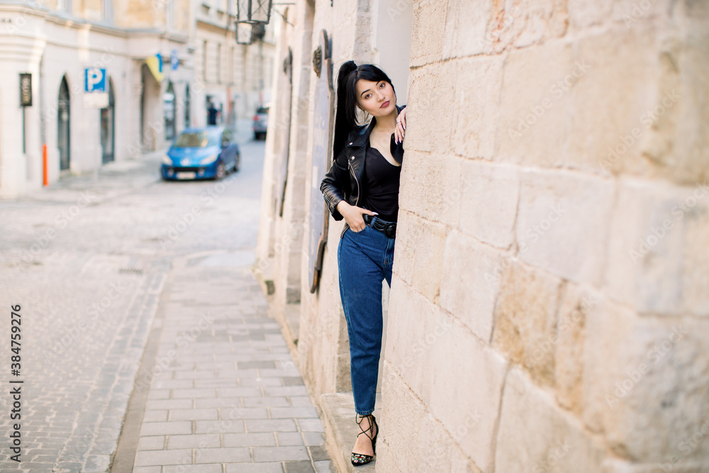 Cute attractive Asian girl in black leather jacket and jeans, posing to camera outdoors in the old city street, hiding her body behind the ancient building wall