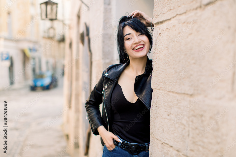 Portrait of stylish young Asian mixed raced woman, wearing black leather jacket, posing to camera, standing near old stone wall of building in ancient European city outdoors