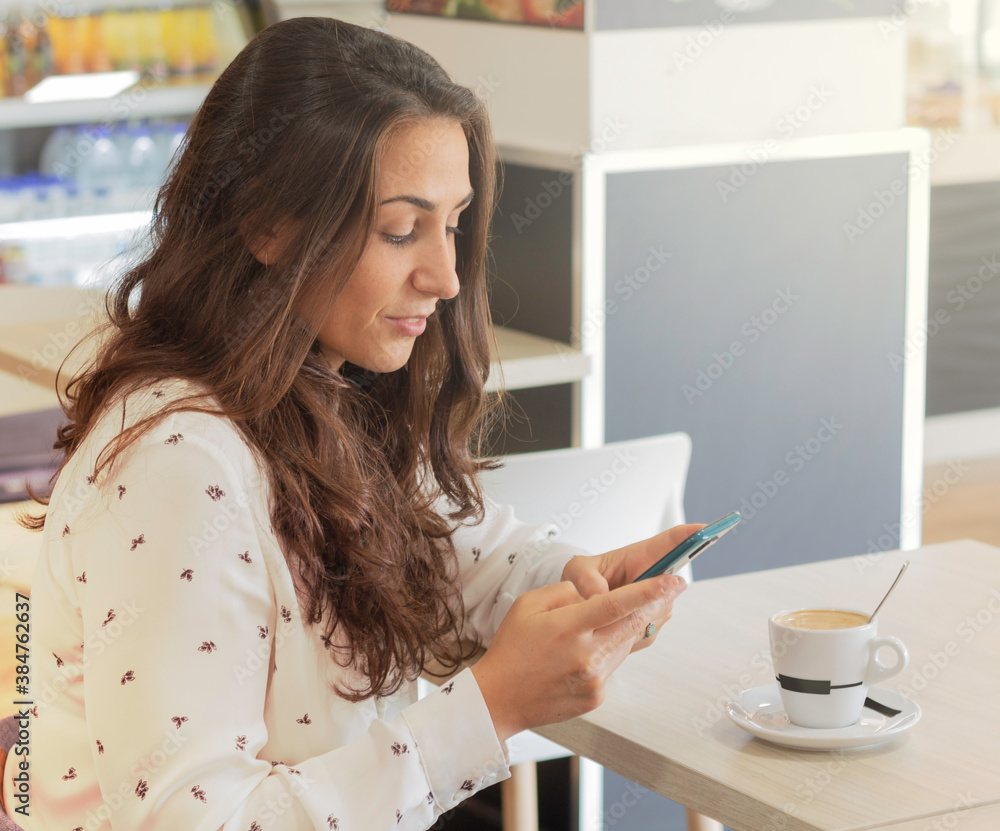 Portrait of smiling young woman using her smartphone in a coffee shop while drinking coffee. Casual business woman using internet with her mobile to telework indoors Portrait of smiling young woman using her smartphone in a coffee shop while drinking coffee. Casual business woman using internet with her mobile to telework indoors