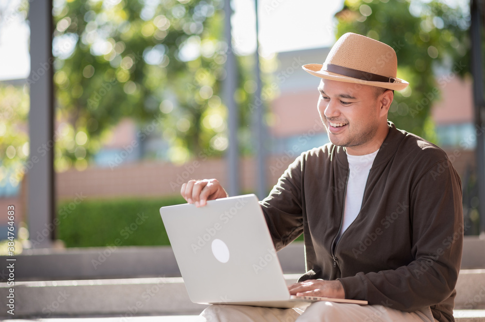 Asian freelancer put on brown hat working outdoor. Office worker or businessman operate on laptop at staircase touch on device enjoy in garden area. Outdoor Activity Concept.