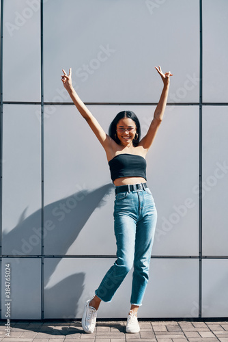 Fototapeta Naklejka Na Ścianę i Meble -  Full length portrait of fashionable young black woman with arms raised showing peace sign
