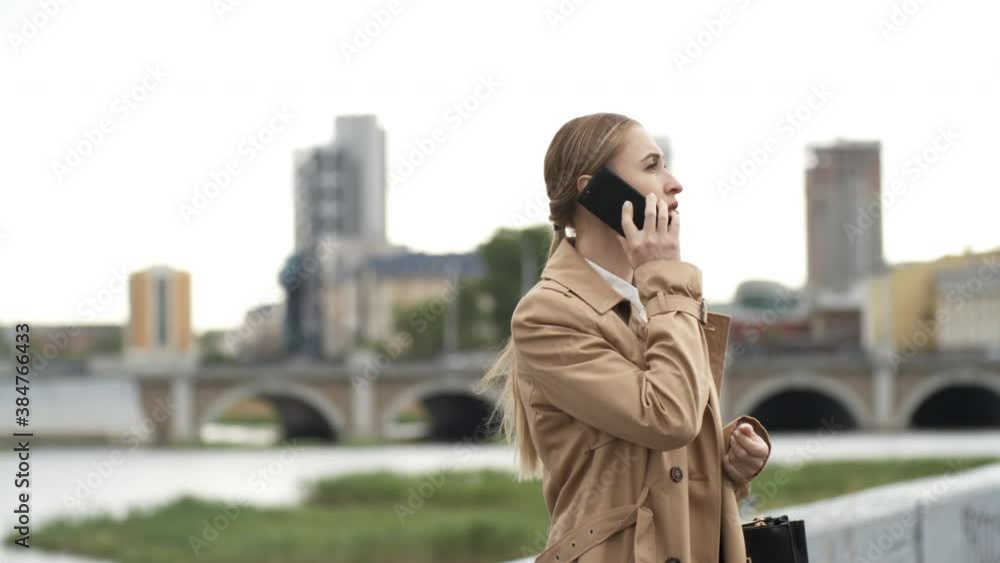 Medium shot of serious businesswoman in trench coat standing on embankment and having phone conversation Medium shot of serious businesswoman in trench coat standing on embankment and having phone conversation