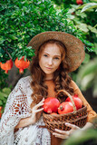Red pomegranate garden with a happy redhead girl in a hat in an orange dress and white shawl. Woman with big red fruits in .fruit basket, with an amazing smile