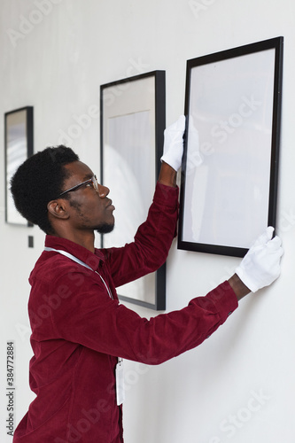 Vertical side view at African-American man hanging frames on wall while planning art gallery or exhibition