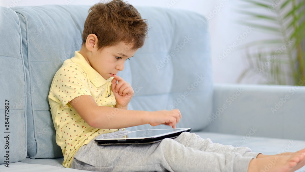 Serious boy watching something interesting from digital tablet sitting on a sofa in the living room at home. Gadgets and technology concept. Serious boy watching something interesting from digital tablet sitting on a sofa in the living room at home. Gadgets and technology concept.