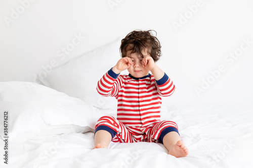 Sleepy toddler wearing pajamas sitting on white bed rubbing eyes