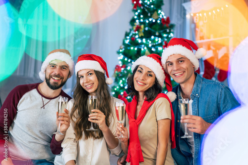 Happy excited group of the best beautiful friends in santa hats with glasses of champagne in hands are clinking while celebrating Christmas holidays