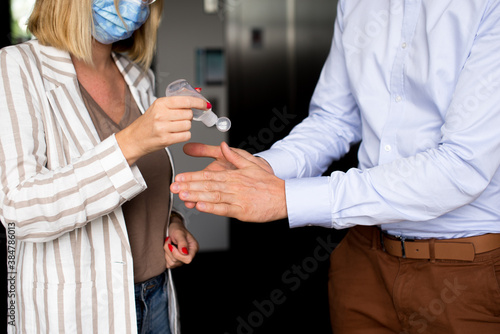 Woman pouring alcohol gel to mans hand. Man and Woman wearing mask. Businessman and businesswoman are washing hands with alcohol gel in office. Coronavirus in corporation. Covid-19 Theme.