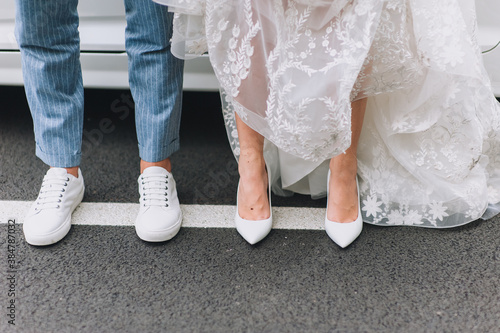 Legs of newlyweds close-up in fashionable white leather shoes on an asphalt road against the background of a car. Photography, concept, copy space.