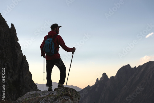 A silhouette of a young man in a cap and sunglasses with a backpack and trekking poles stands high in the mountains in the evening against the backdrop of the ridge.
