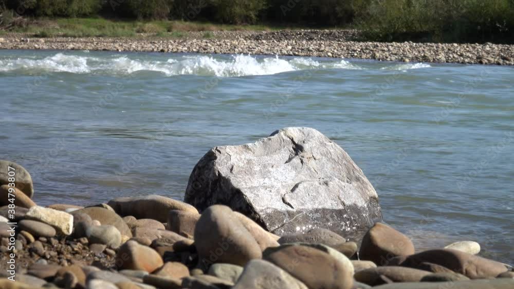 Stones and pebbles on the mountain river bank against the background of a fast-flowing river Stones and pebbles on the mountain river bank against the background of a fast-flowing river