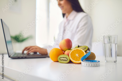 Fruit, measuring tape and glass of water placed on desk against blurred dietitian working on laptop