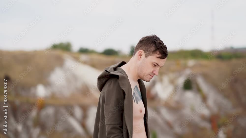 Tattooed man with bare torso in cape looks around thoughtfully in cool weather on walk. Adult pondering male stands on windy day in hilly terrain. Slow motion. Tattooed man with bare torso in cape looks around thoughtfully in cool weather on walk. Adult pondering male stands on windy day in hilly terrain. Slow motion.