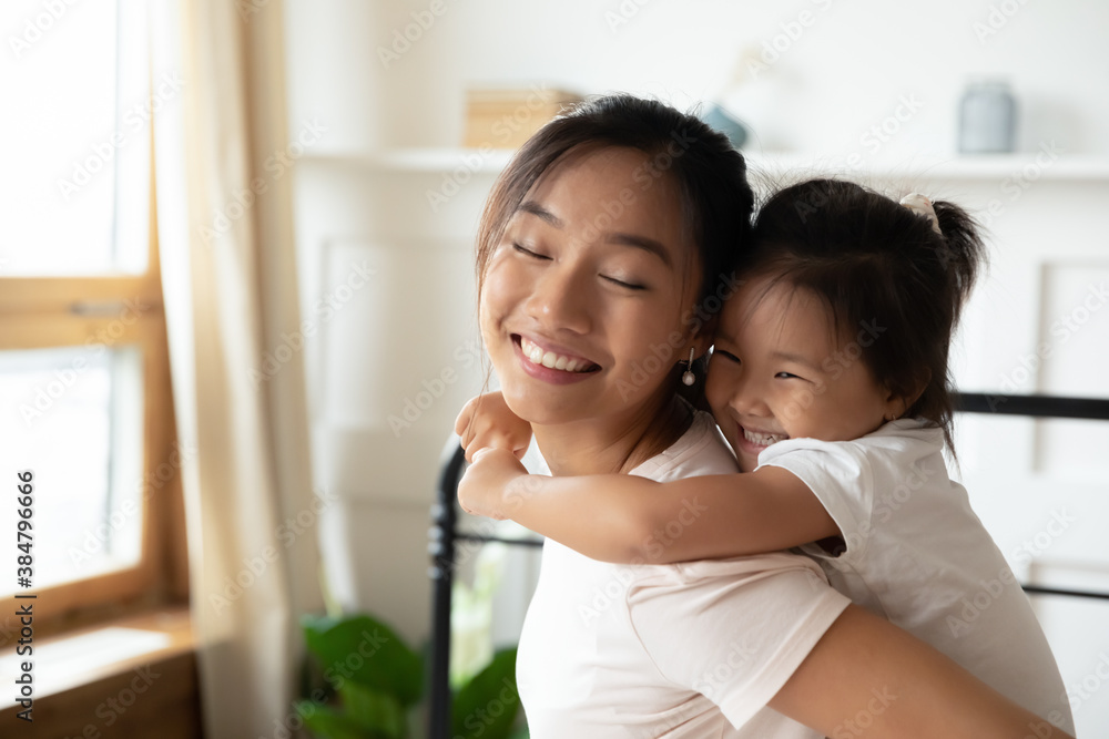 Close up smiling Asian mother piggy backing adorable little daughter, standing in bedroom, happy loving young mum and cute toddler girl kid enjoying tender moment together, cuddling hugging Close up smiling Asian mother piggy backing adorable little daughter, standing in bedroom, happy loving young mum and cute toddler girl kid enjoying tender moment together, cuddling hugging