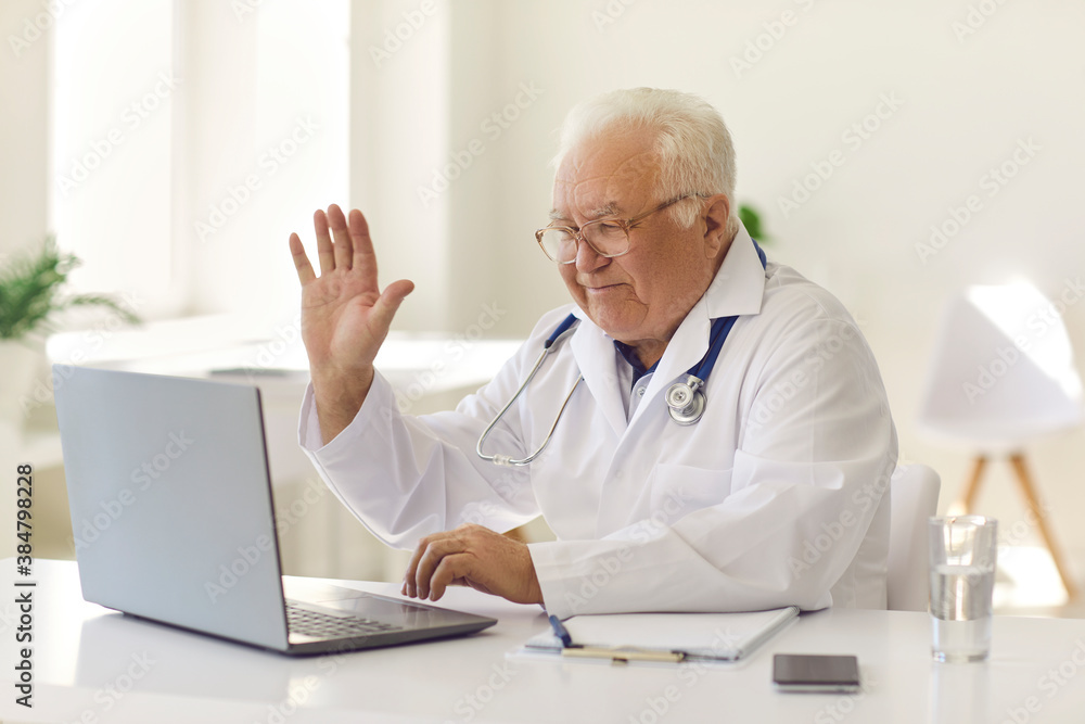 Friendly senior doctor waving hand at laptop screen starting online consultation with patient