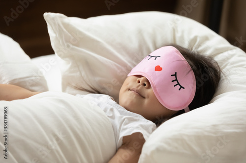 Close up cute Asian little girl wearing pink mask sleeping, taking nap in cozy bed alone, tranquil pretty toddler child resting on soft pillow under warm blanket duvet in bedroom at home photo