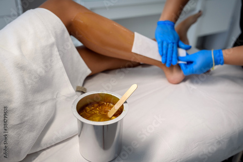 A master applies pink depilatory wax to a young woman's leg for hair removal. Depilation with wax. Beauty concept. Place for text. Selective focus.