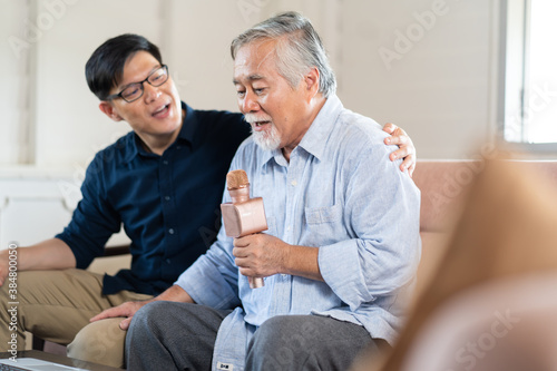 Happy Asian family. Senior Asian father and Adult son singing karaoke on the sofa in the living room at home. Home Entertainment.