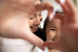 Close up smiling Asian mother and little daughter making heart gesture, looking at camera through fingers, loving young mum and adorable toddler girl showing love sign, posing for selfie photo