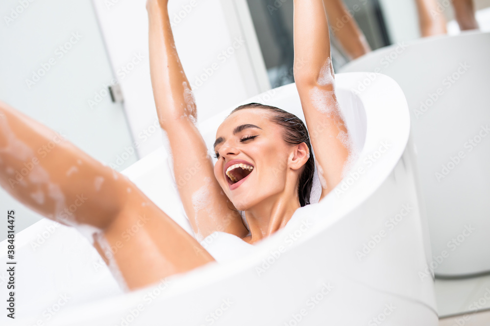 Obraz premium Happy young woman shaving her legs and having fun while lying in bathtub
