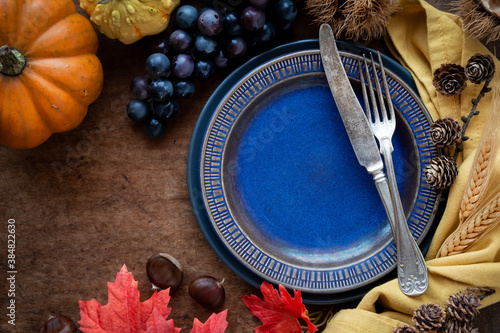 Thanksgiving table setting with plates, vintage silverware and traditional autumnal decorations like pumpkin, chestnuts, grapes, corncobs and pinecones on dark rustic wooden table photo